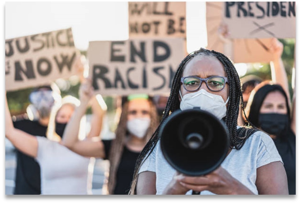 People in masks at a rally. A black woman holds a megaphone, other people in the background hold signs with phrases such as 'End Racism'.